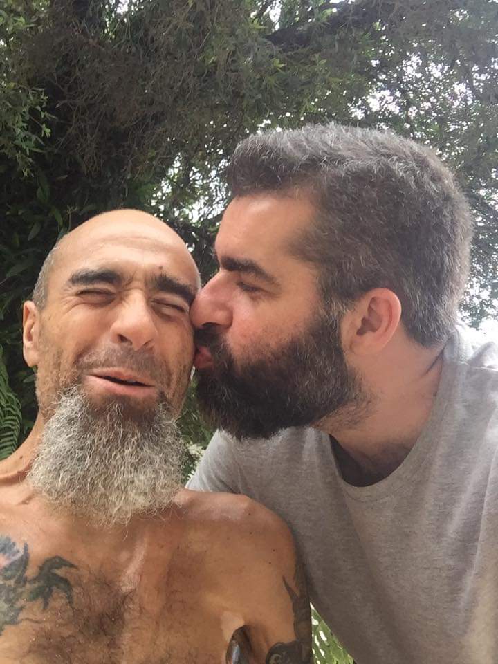 Foto de Renato, um homem sem cabelos e uma longa barba grisalha recebendo um beijo na bochecha de outro homem, de cabelos e barba escuros. Ele está sorrindo de olhos fechados