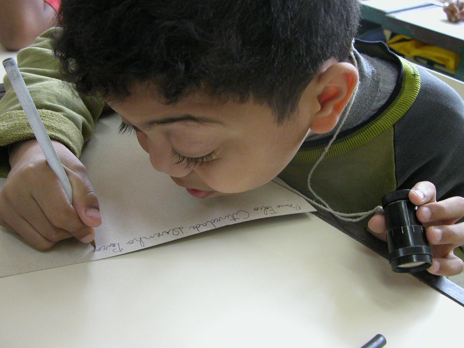Criança com baixa-visão segura tele-lupa com uma das mãos e com a outra um lápis.