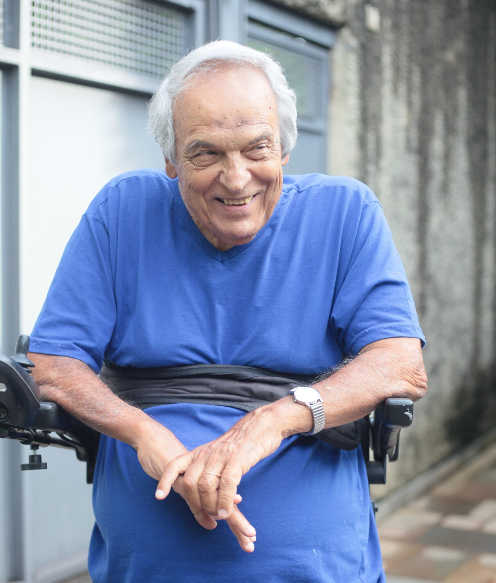 João Carlos Pecci, homem branco com cabelo grisalho. Está em pé com cadeira stand, em sua casa, em São Paulo. Olha para o lado e sorri.