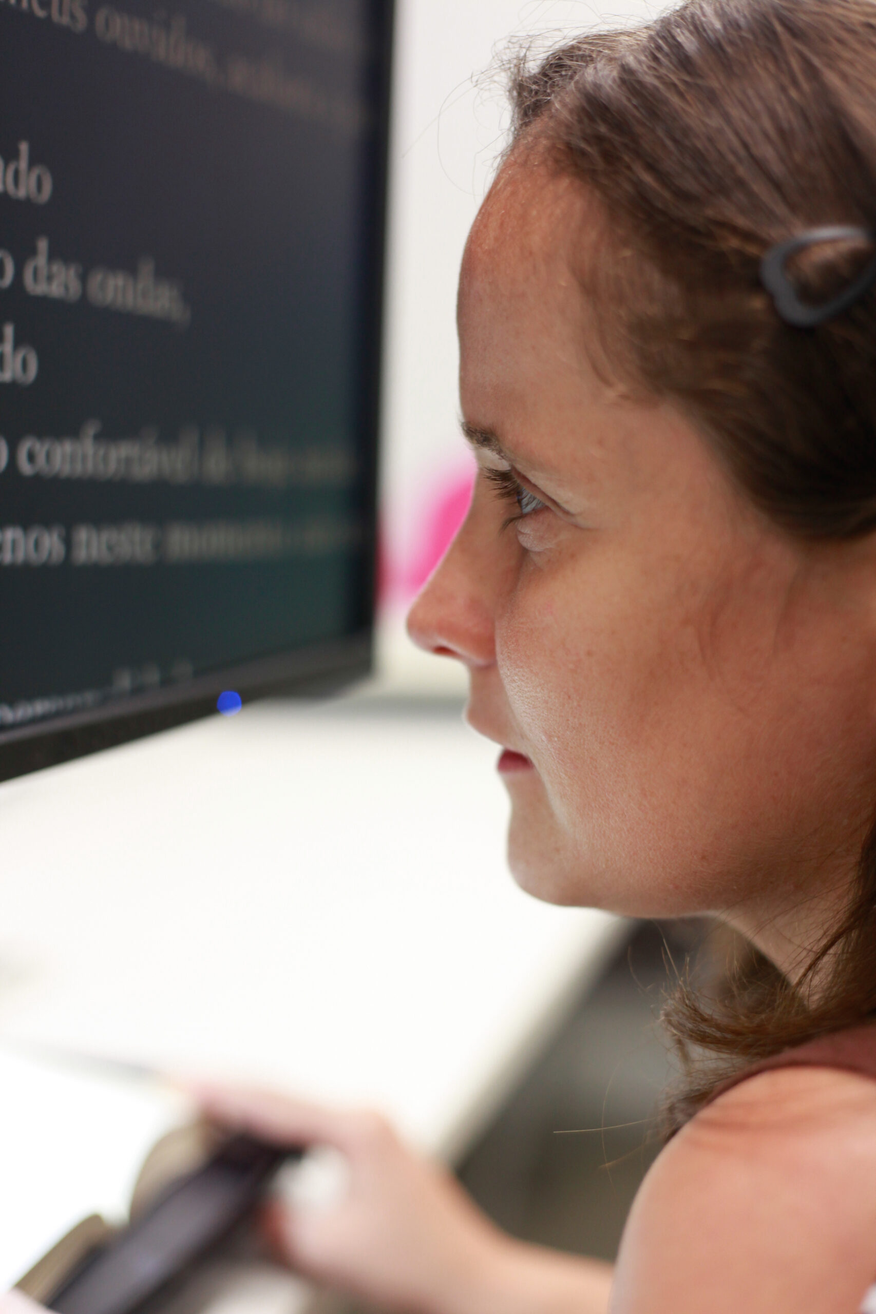 Fotografia colorida de mulher, pele clara, cabelo cacheado e com baixa visão está com o rosto próximo a tela de uma computador. Ela usa recursos de acessibilidade em página da internet. O fundo da tela está escuro e as letras do texto na cor branca e em tamanho grande. Fim da descrição.