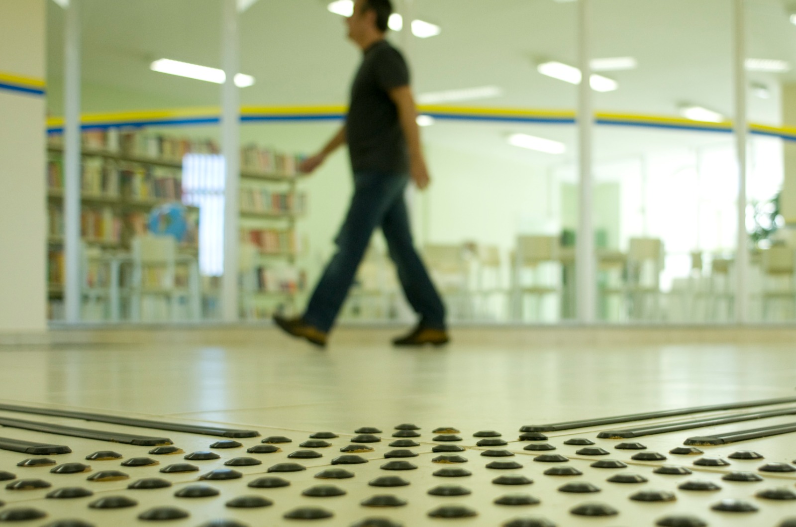 Em foco no primeiro plano, piso tátil preto sobre chão claro. Ao fundo, desfocado, um homem caminha pelo corredor.