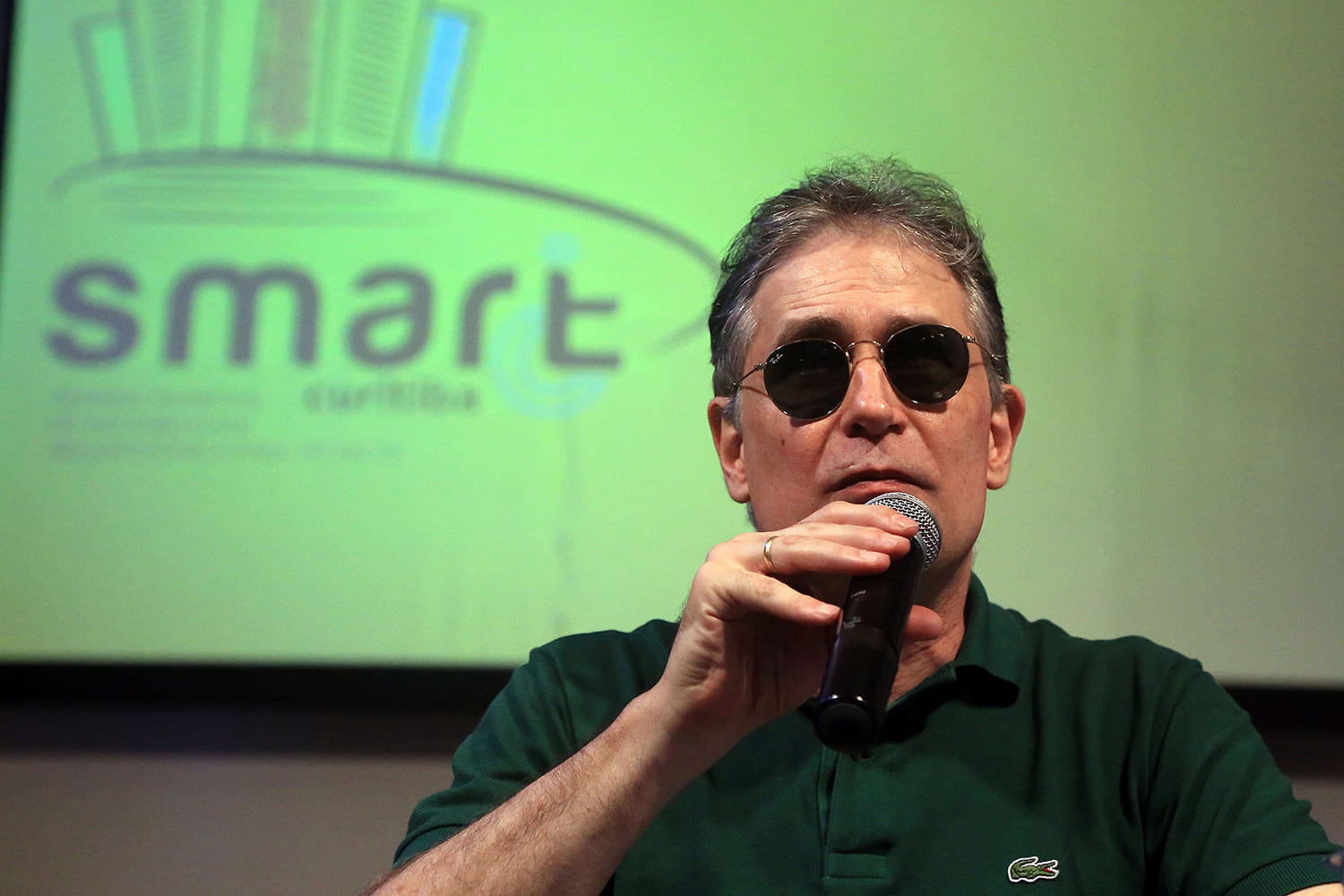 Desembargador cego, Ricardo Tadeu está segurando o microfone. Usa óculos escuros e camisa verde. Atrás há um telão com o nome Smart, evento sobre acessibilidade arquitetônica para técnicos.