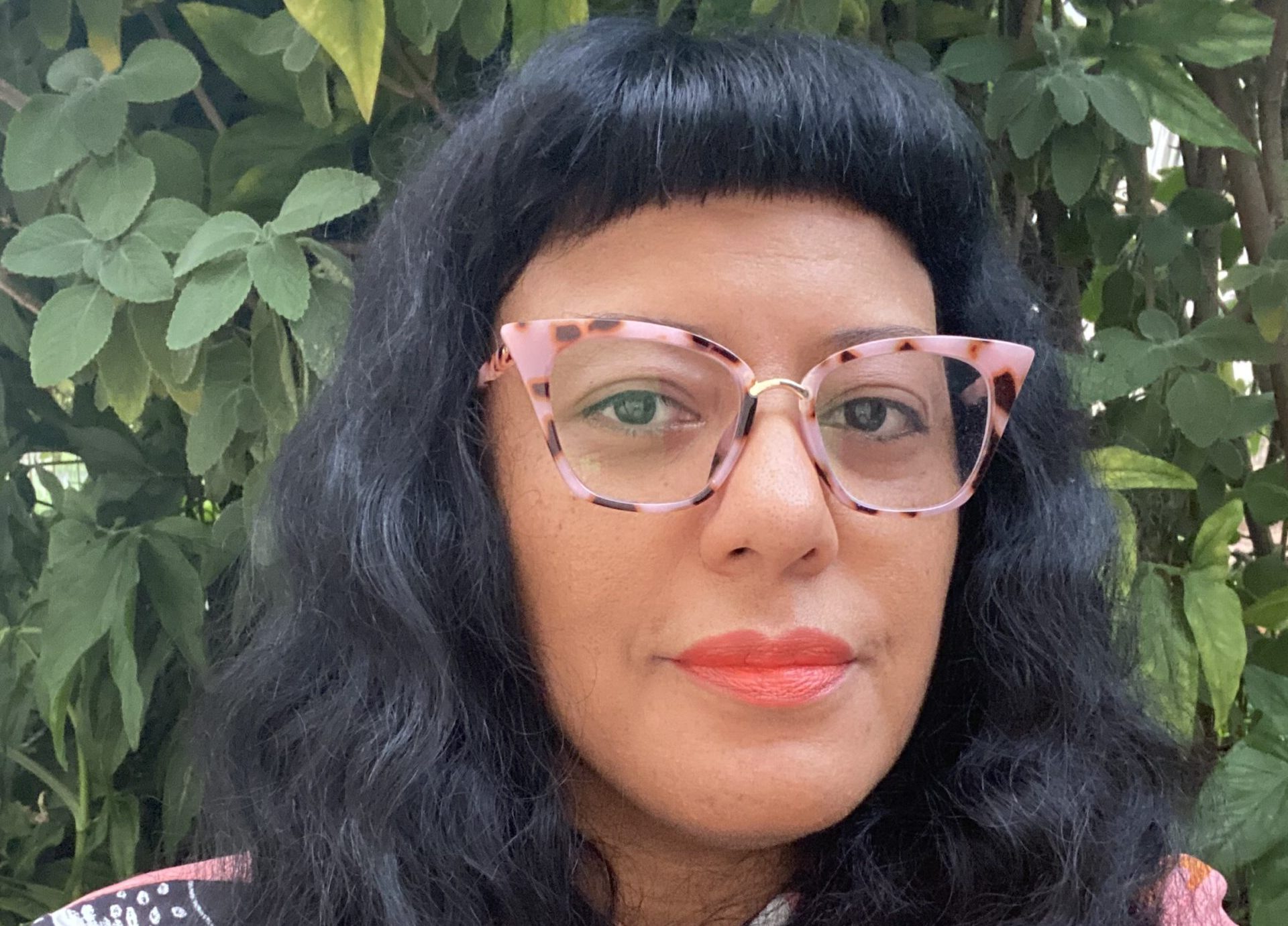 Foto de Andreia Freitas, uma mulher negra, de cabelos longos e caccheados com uma franja e óculos com armação rosa. Ela olha na direção da câmera e há plantas ao fundo