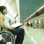 Homem branco em cadeira de rodas, com cabelo rastafári, lendo livro na estação de trem.