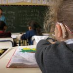 Foto em sala de aula, com foco na cabeça de criança com aparelho auditivo. Em segundo plano, lousa com anotações, colegas de classe e professora.