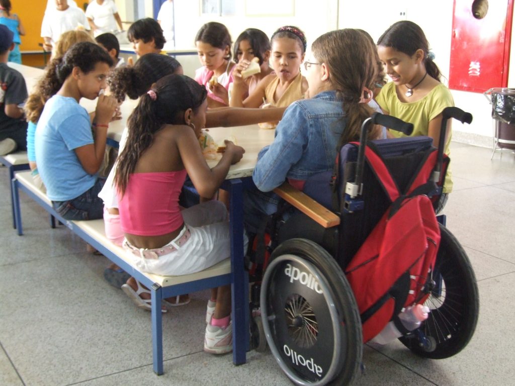 Crianças na mesa do recreio, em escola, uma delas em cadeira de rodas.