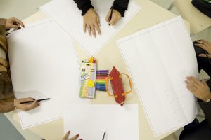 Foto de mesa vista de cima, com papéis, caixa de lápis de cor e mãos adultas escrevendo nas folhas espalhadas.