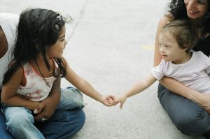 Foto de duas meninas pequenas sorrindo tocando as mãos uma da outra. A menina no lado direito tem Síndrome de Down
