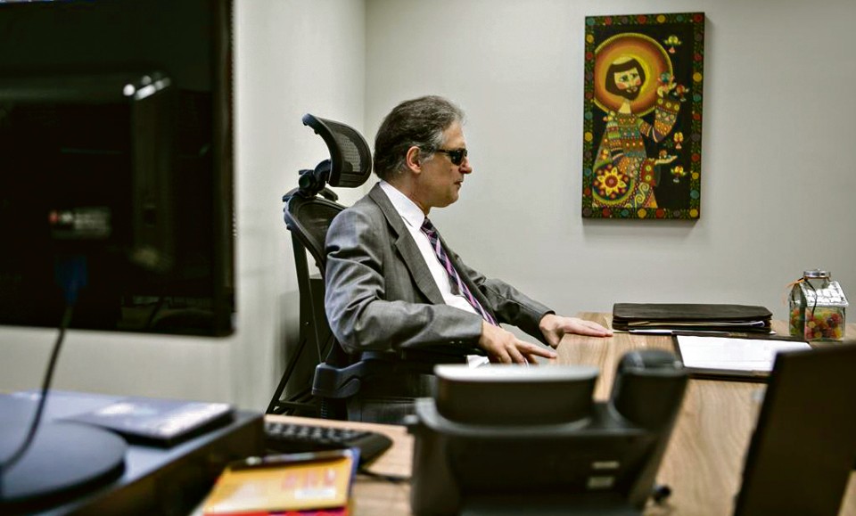 Ricardo Tadeu, desembargador cego, em ambiente de trabalho. Está sentado à mesa, usa óculos escuros, terno e gravata. Ao lado, parede com um quadro.