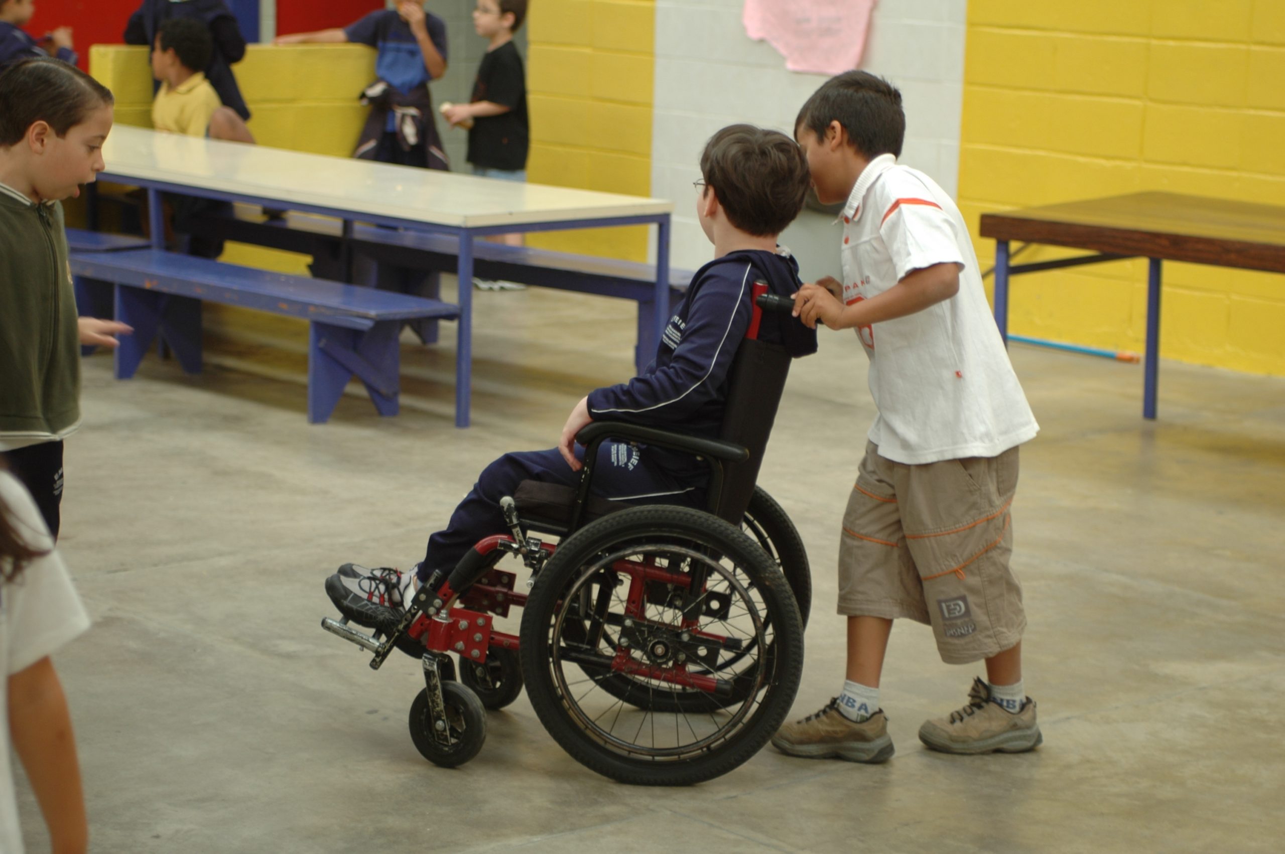 Aluno empurra a cadeira de rodas do colega em um pátio escolar com outras crianças.