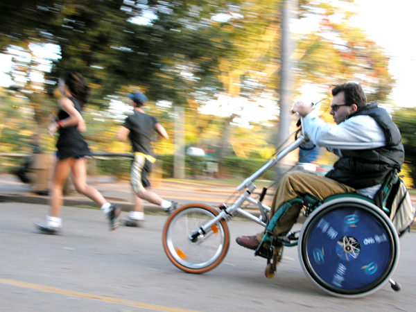 Foto de um homem cadeirante em uma bicicleta adaptada, ele está em um espaço aberto e arborizado, com pessoas praticando atividades físicas.