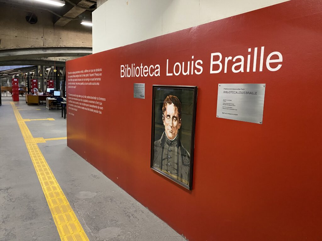 Foto de um dos corredores da Biblioteca Louis Braille, com um retrato de Louis Braille pendurado em uma parede vermelha