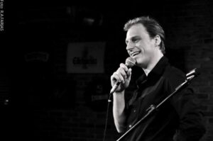 Foto em preto e branco de Fagner Zadra, um homem branco de cabelos curtos e escuros em um palco, segurando um microfone e olhando para a esquerda