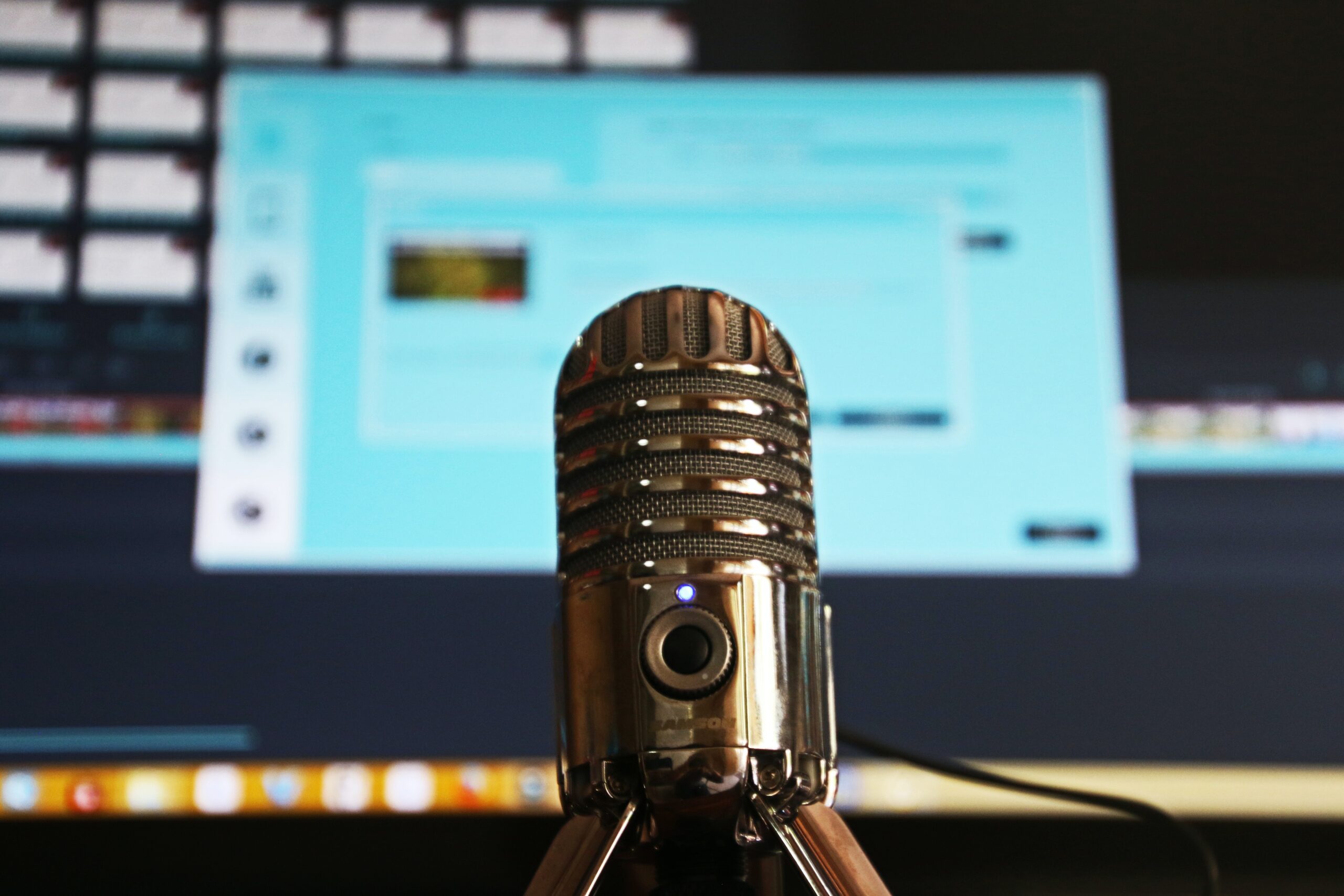 Foto em close de um microfone de mesa, com uma tela de computador desfocada ao fundo
