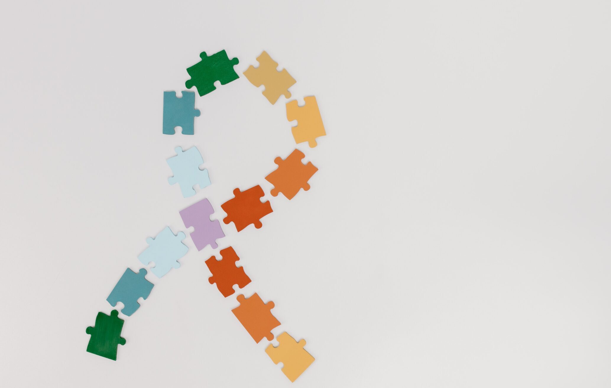 Foto em fundo branco, com peças de quebra-cabeça coloridas foramando um laço, símbolo do autismo