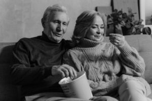 Foto em preto e branco de um casal de idosos sentado em um sofá, sorrindo e comendo pipoca, próximos um do outro