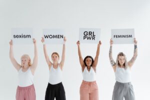 Foto com um grupo de quatro mulheres jovens, de diferentes etnias, erguendo cartazes. Texto dos cartazes, da esquerda para a direita: "sexism", com um risco em cima, "woman", Girl Power" e "Feminism"