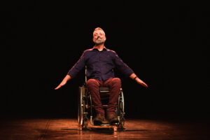 Foto do ator Luciano Mallmann em um palco, com fundo escuro. Ele é branco, com cabelos grisalhos e está em uma cadeira de rodas, no centro, com os braços abertos para baixo