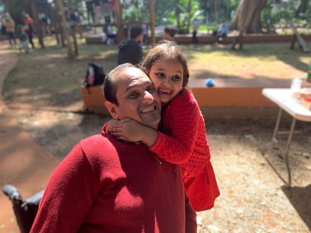 Foto de Jairo Marques com a filha Elis, abraçados e sorrindo. Ele é um homem pardo, de cabelos curtos e usa uma camisa vermelha. Elis tem cabelos ondulados e usa um casaco vermelho