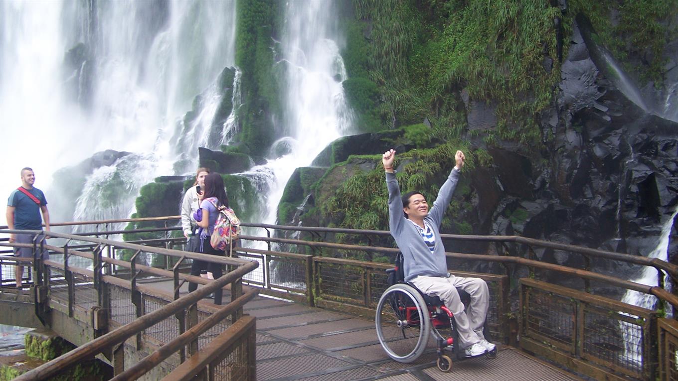 Foto de Ricardo em meio à uma ponte próximo à uma cachoeira, em uma cadeira de rodas com os braços erguidos para o alto