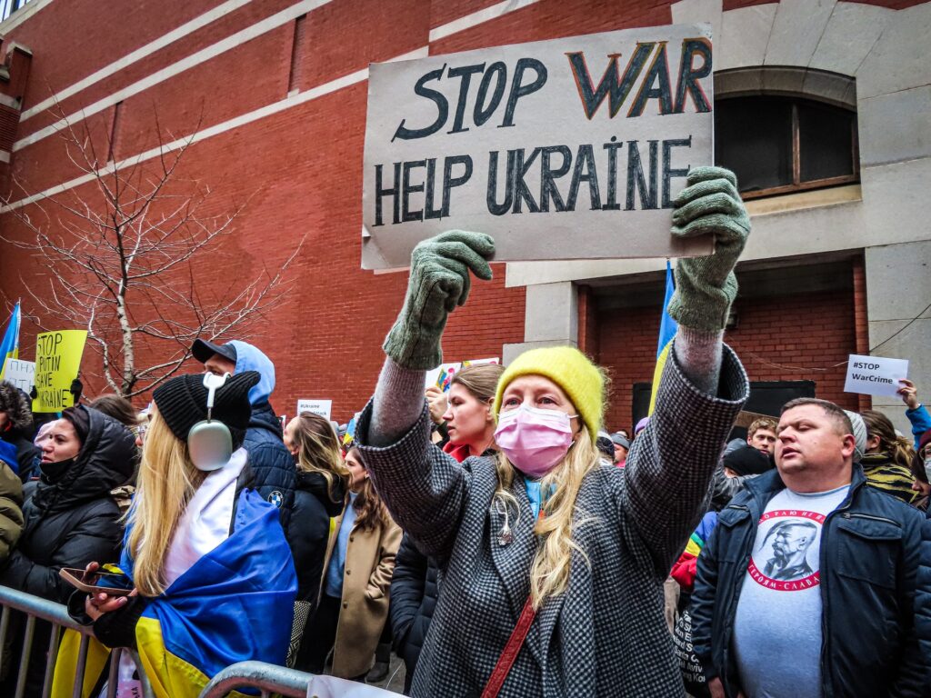 Foto de um grupo de pessoas na rua, de máscaras, protestando. Uma mulher loira segura um cartaz com o texto: Stop war help Ukraine