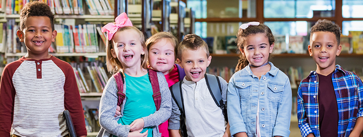 Foto com um grupo de seis crianças, três meninas e três meninos, sorrindo e olhando na direção da câmera. Elas estão em uma sala com estantes de livros ao fundo
