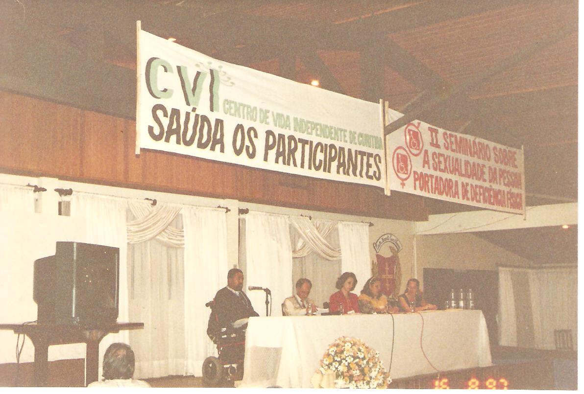 Foto de um auditório, com mesa no palco e cinco pessoas.