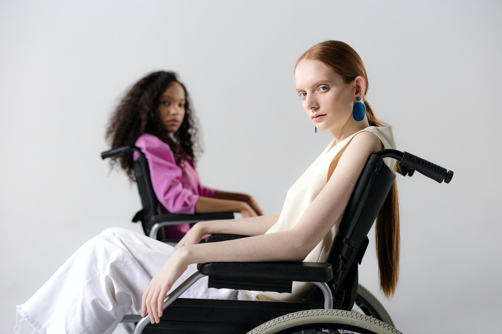 Foto de duas mulheres jovens, uma branca e uma negra, de lado, em cadeiras de rodas. Elas olham em direção à câmera e estão com expressões sérias